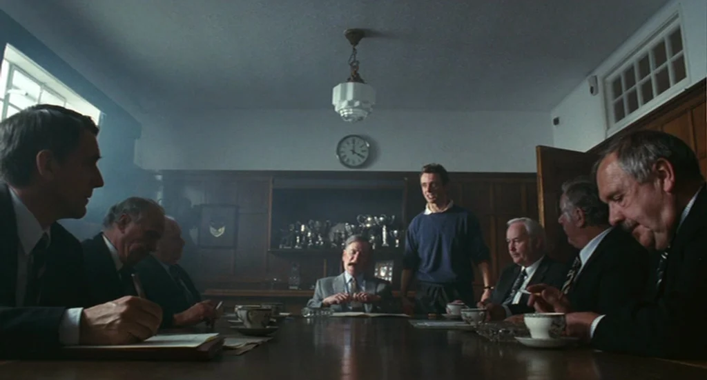 The Damned United (2009) movie still: wide — A dark, wood-paneled room features a central clock and trophy shelf on the back wall. A group of men in…