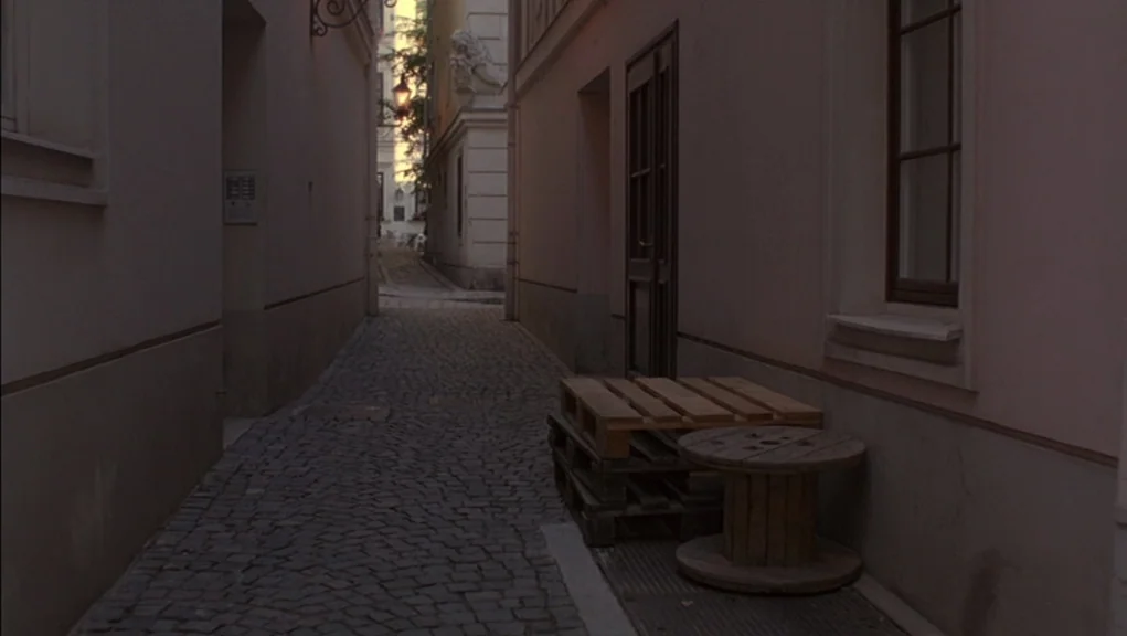 Before Sunrise (1995) movie still: establishing — A narrow cobblestone alleyway sits between two beige-colored stucco buildings. A stack of wooden…