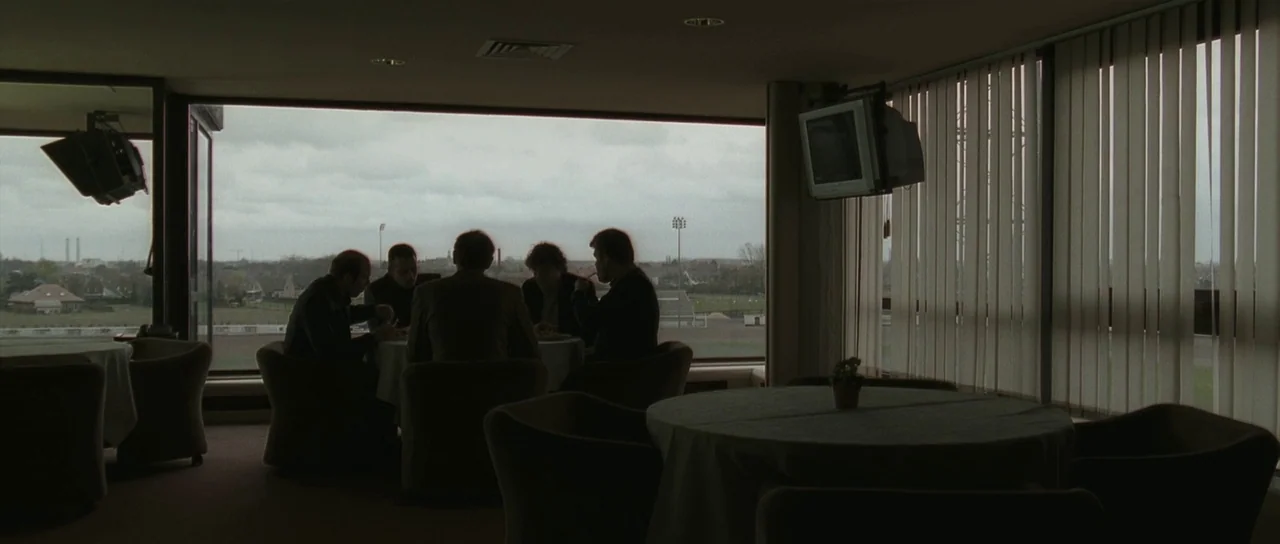 Bullhead (2011) movie still: wide — A dimly lit room with large windows overlooking a distant field and grey sky. Five men are seated around a…