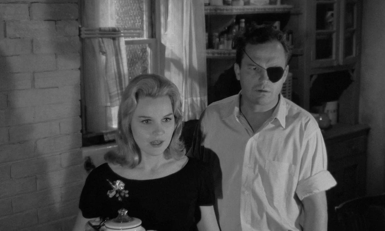 Something Wild (1961) movie still: medium — A domestic space with a brick wall and a window draped in light curtains. A woman and a man wearing an…