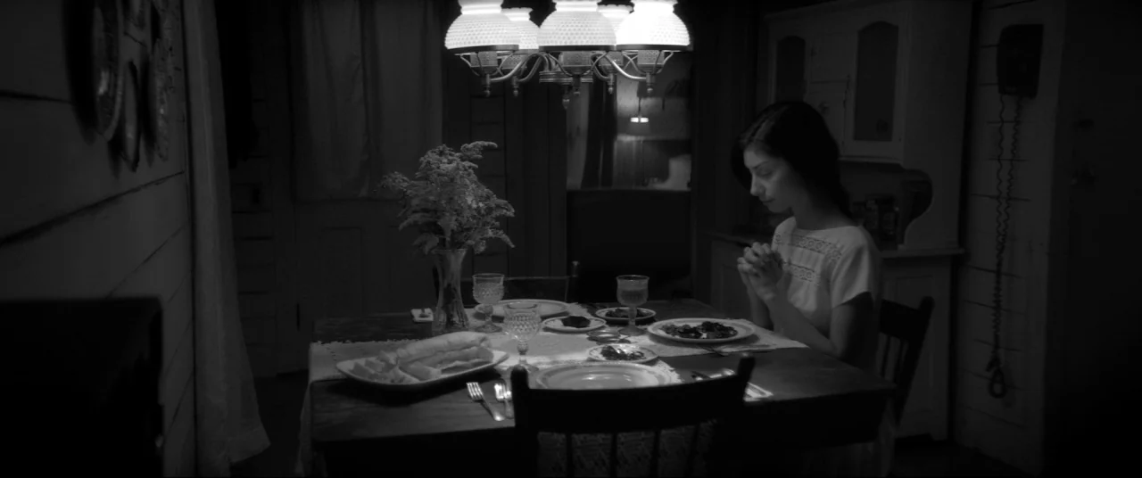 The Eyes of My Mother (2016) movie still: medium — A dimly lit room features a wooden table set for dinner with floral arrangements. A young woman…