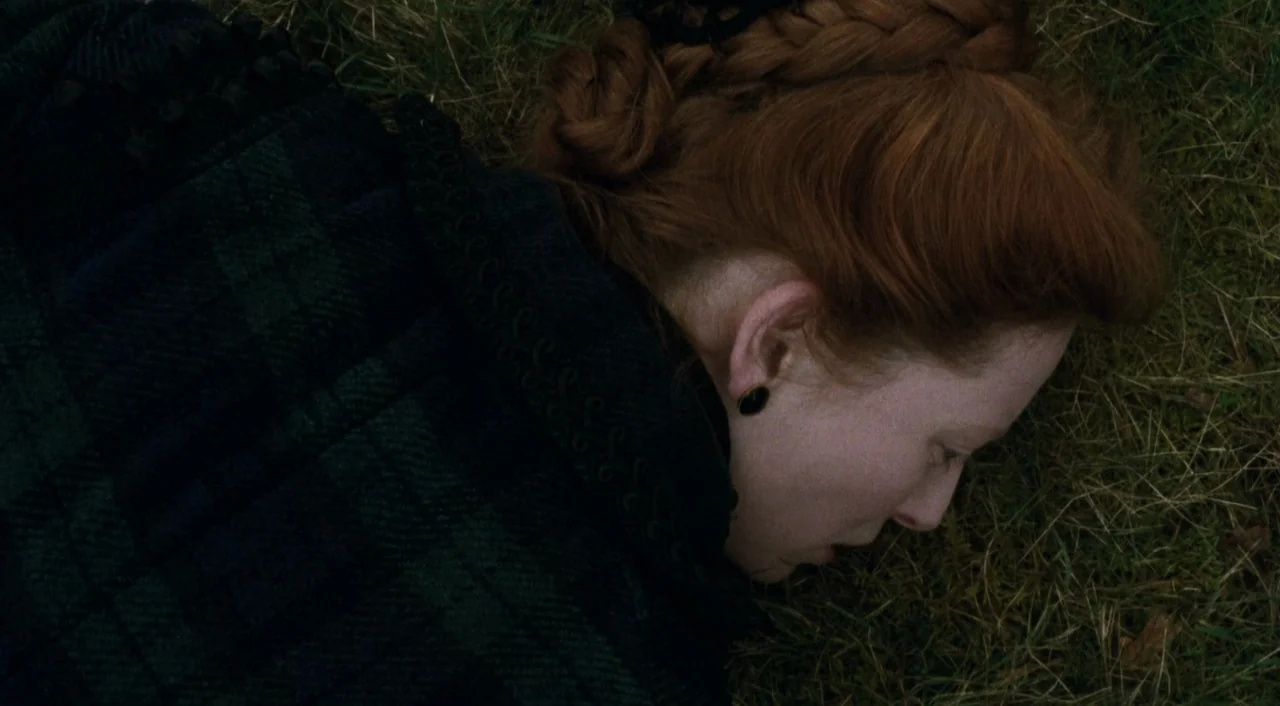 Orlando (1992) movie still: closeup — A woman lies face down in a patch of dry green grass. She wears a thick plaid coat with a complex braided…