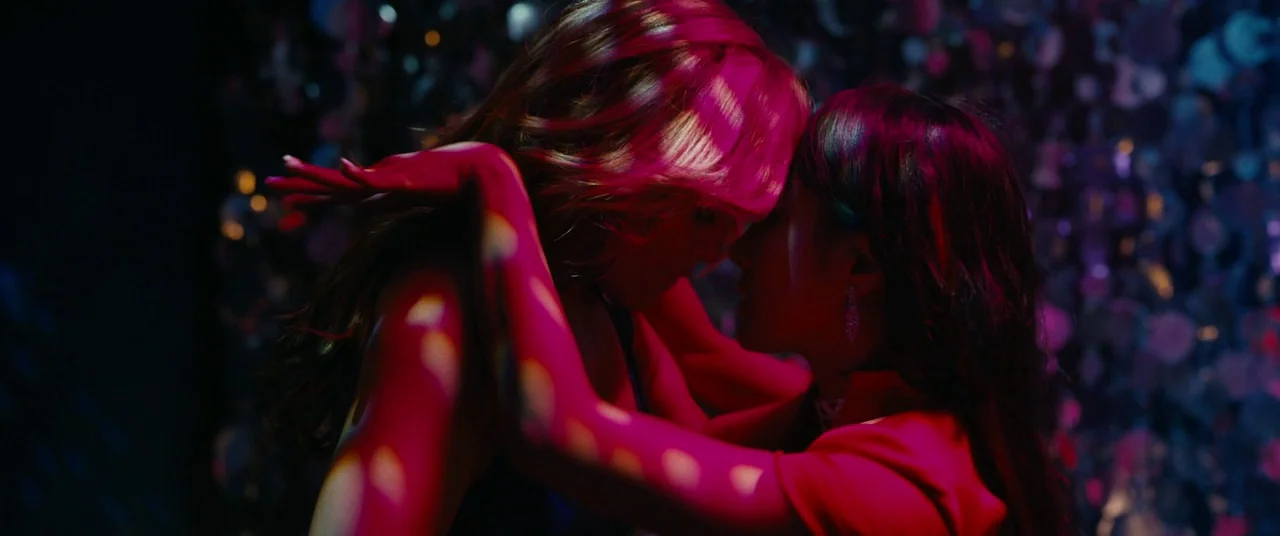 Hustlers (2019) movie still: medium — A dark, dimly lit environment features a shimmering, textured background surface. Two women embrace closely…