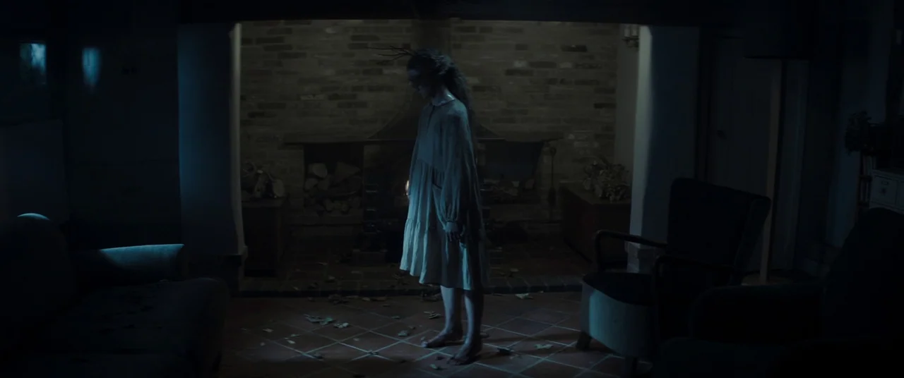 Lord of Misrule (2023) movie still: fullbody — A dimly lit room features a stone fireplace with scattered dried leaves on tiles. A young girl stands…