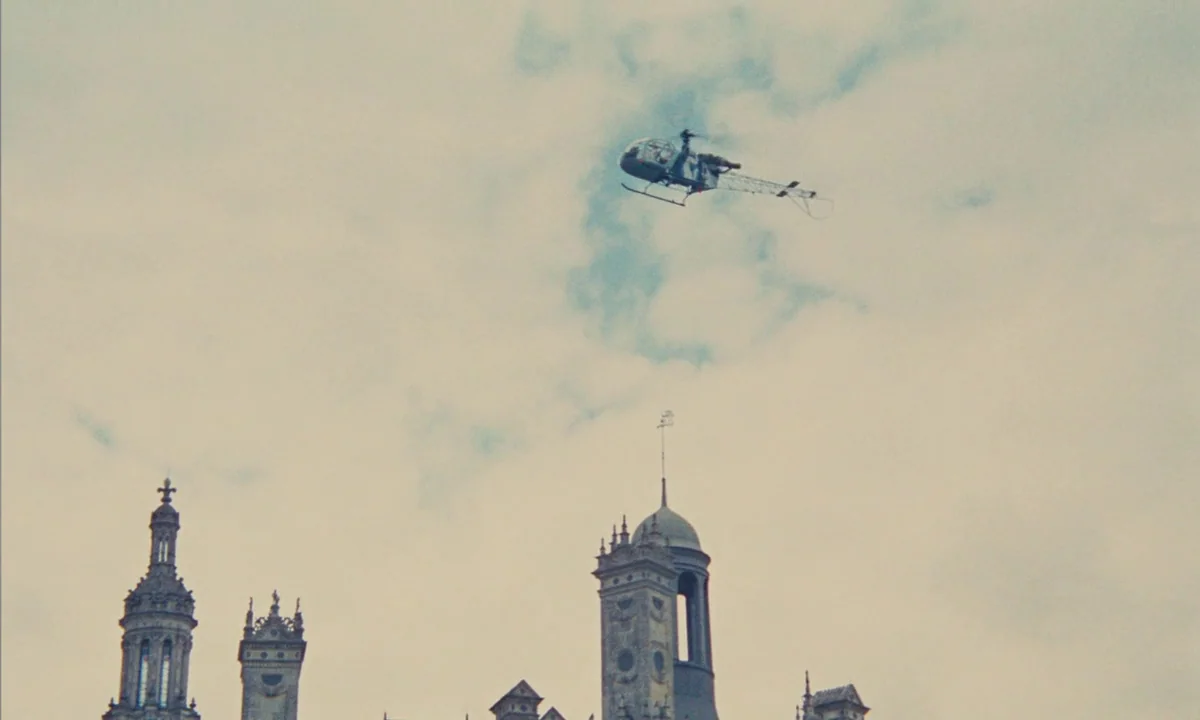 Donkey Skin (1970) movie still: wide — An ornate historic chateau with multiple towers sits beneath a cloudy, overcast sky. A small helicopter…