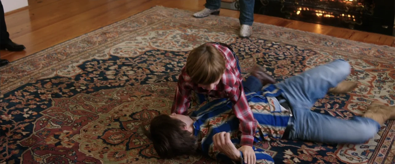 Vacation (2015) movie still: medium — A residential room features a large, detailed oriental carpet covering the hardwood floor. Two people wrestle…