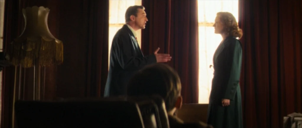 Nowhere Boy movie still: medium — A dimly lit room features heavy dark red curtains covering a bright window. A man in a suit faces a woman in a…