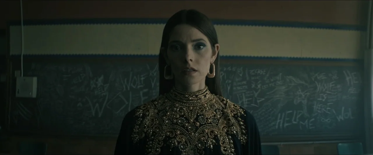 It Feeds (2025) movie still: medium — A dark classroom features a chalkboard covered in white chalk scribbles and urgent text. A woman stands…