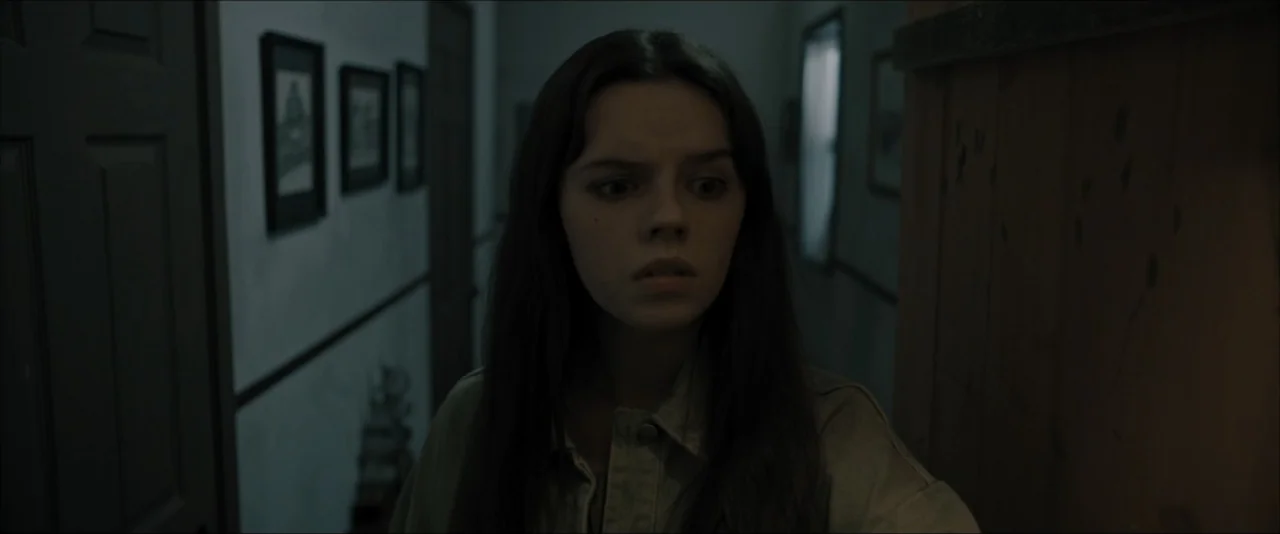 It Feeds (2025) movie still: medium — A dimly lit indoor residential corridor with framed pictures on the wall. A young woman with long dark hair…