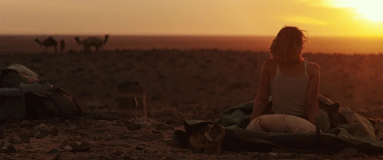 Tracks (2013) movie still: wide — A vast rocky desert expanse is visible under a vibrant sunset sky. A young woman sits on the ground wrapped in a…