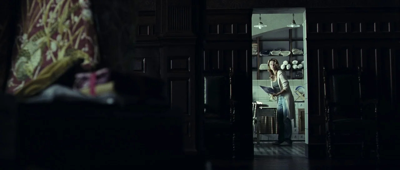 The Orphanage (2007) movie still: wide — A woman stands in a bright, illuminated room viewed through a dark wooden doorway. She holds a paper and…