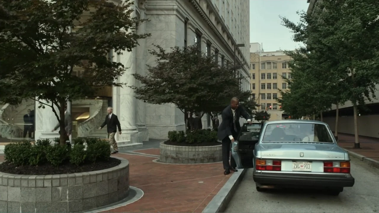 Confirmation (2016) movie still: wide — A city street features a large marble building with a circular planter and trees. A man in a dark suit steps…