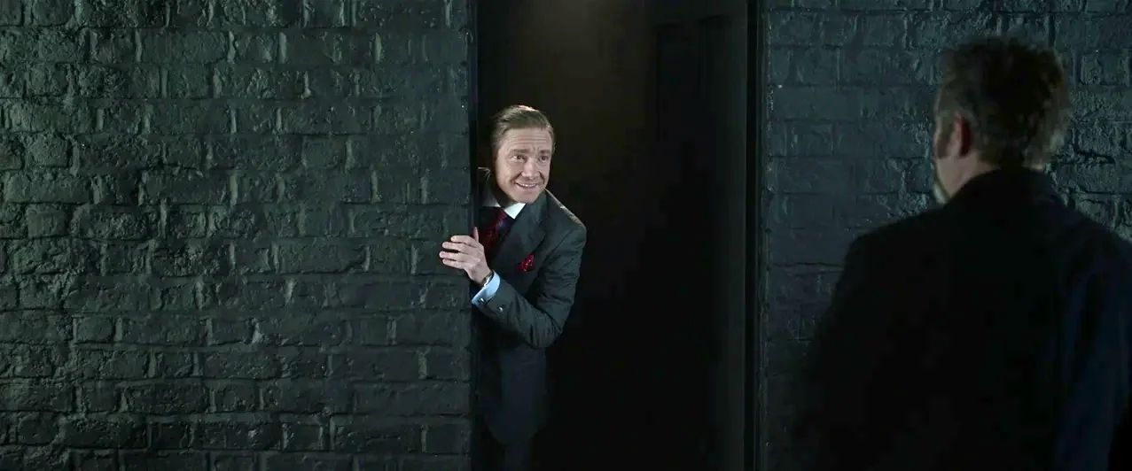 Ghost Stories (2017) movie still: medium — A dark, dimly lit space features a painted black brick wall and an open door. A man in a suit peeks…