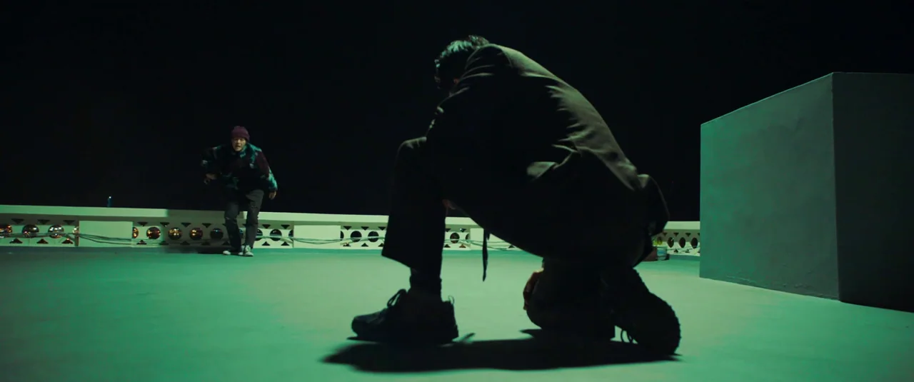 Decision To Leave (2022) movie still: fullbody — A wide, flat rooftop at night features a low perimeter wall with circular cutouts. One man kneels…