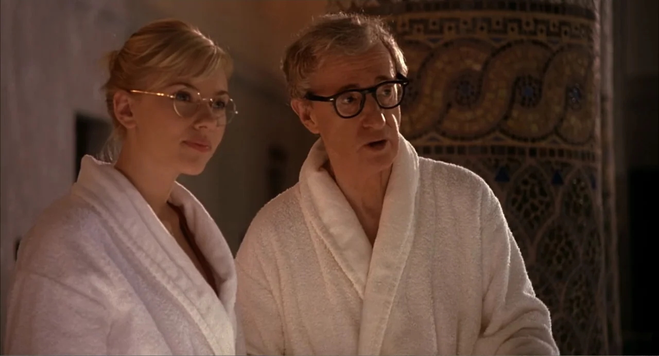 Scoop (2024) movie still: medium — Two individuals stand in a room featuring a tiled mosaic column nearby. Both people wear white bathrobes and…