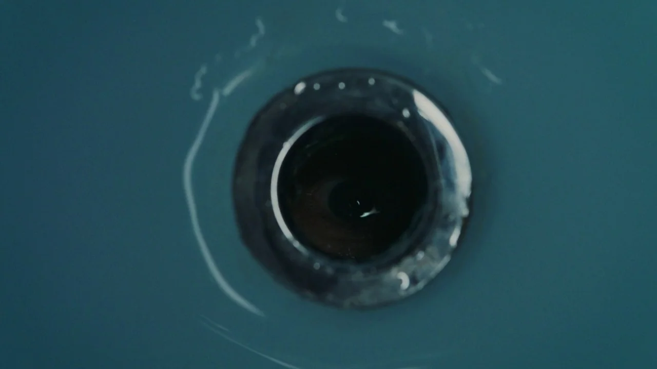 Girl on the Third Floor (2019) movie still: closeup — A close-up view of a metal sink drain surrounded by a matte blue basin. An eye is visible…