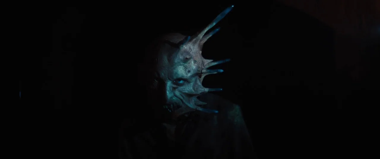 Daniel Isn’t Real movie still: closeup — A dark, featureless space hides the surroundings in deep shadow. A monster with sharp facial spikes stares…