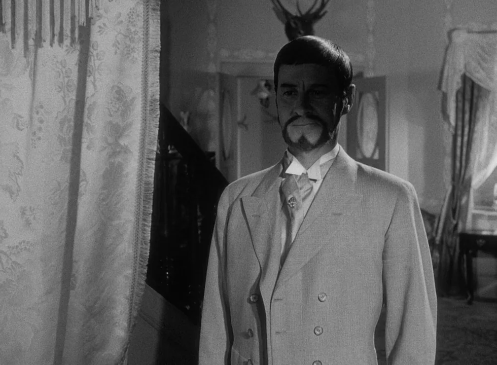 Smiles Of A Summer Night (1955) movie still: medium — A furnished parlor contains floral curtains, a dark staircase, and an antler wall mount. A man…