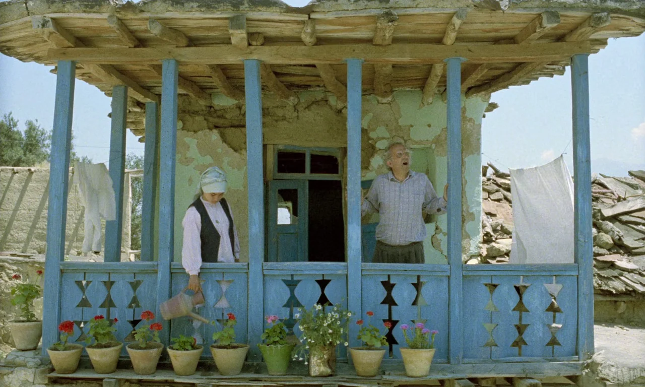 Life, and Nothing More (1992) movie still: fullbody — A rural wooden porch with blue railings and many potted plants in clay pots. A young woman…