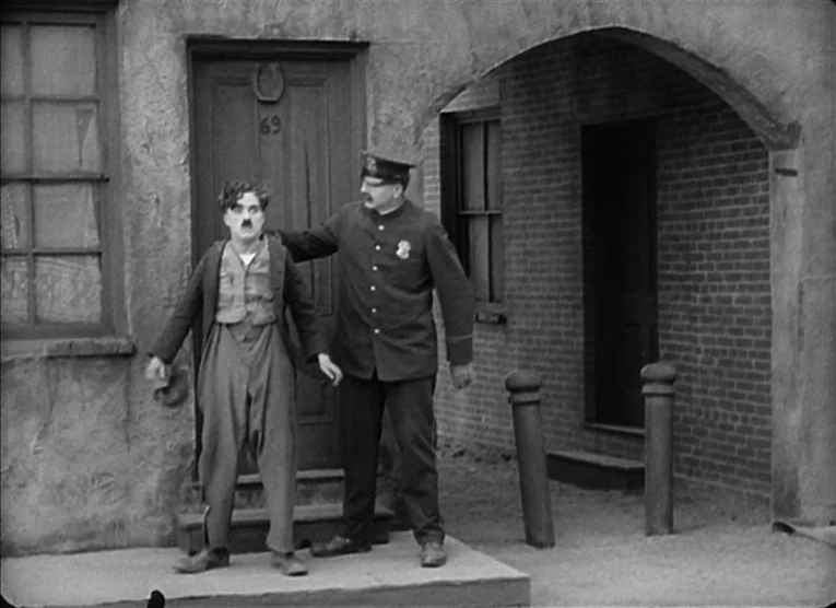 The Kid (1921) movie still: fullbody — Two men stand before a dark wooden door with a horseshoe above the frame. A police officer holds the shoulder…