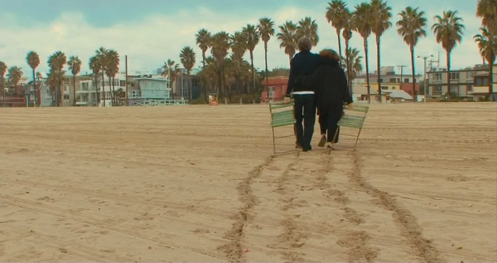The Beaches of Agnès (2008) movie still: wide — A wide view of a sandy coastline lined with tall palm trees and houses. Two people walk away from…