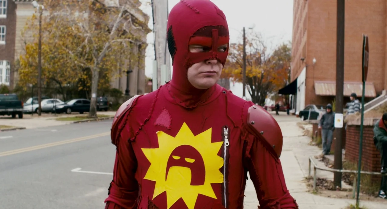 Super (2010) movie still: medium — A man dressed in a homemade superhero costume stands on an urban street corner. Behind him, parked vehicles line…