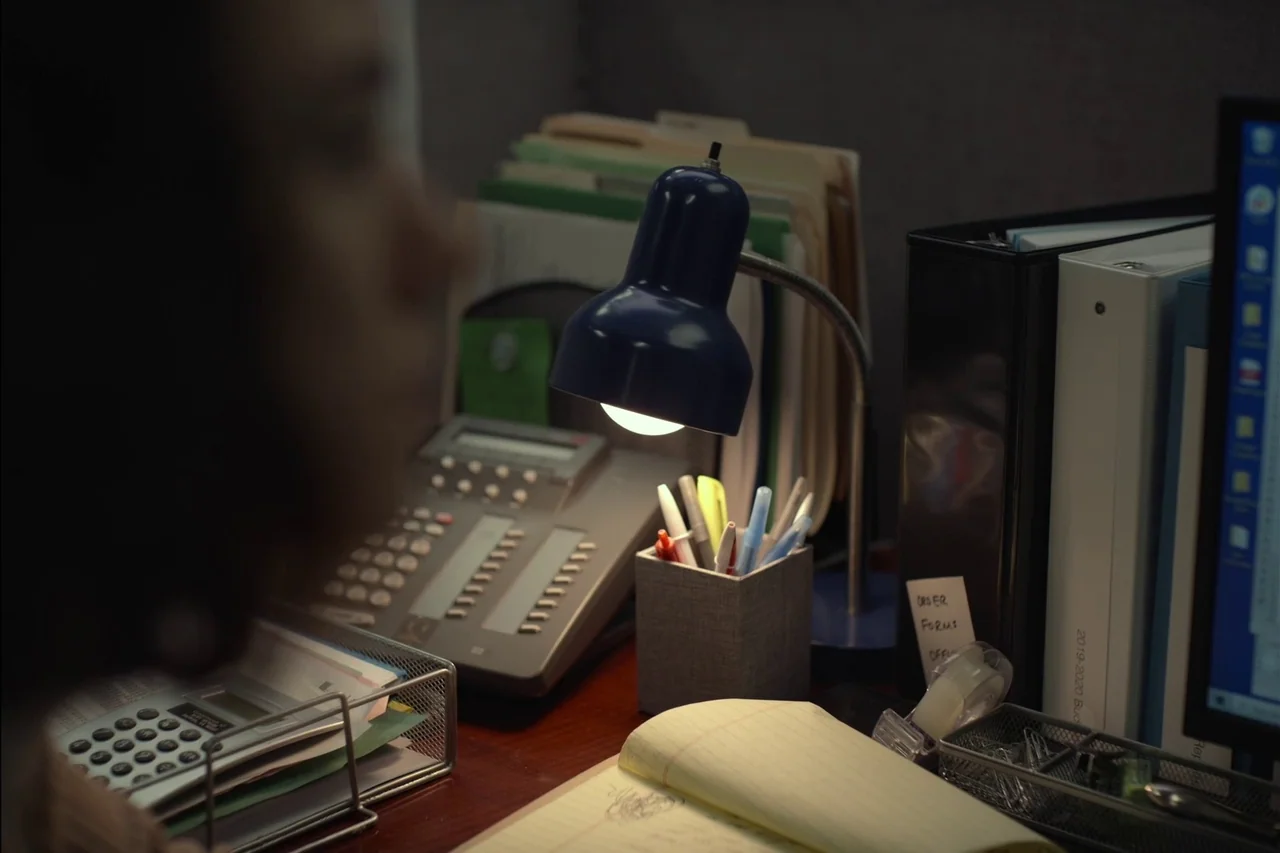 Sometimes I Think About Dying (2023) movie still: medium — An office cubicle desk contains a blue lamp, telephone, monitor, and organizers. A person…