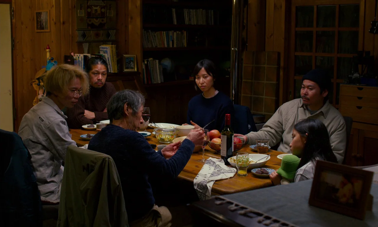 Evil Does Not Exist (2023) movie still: wide — A wooden interior room with bookshelves and paneled walls surrounds a dining table. Six family…