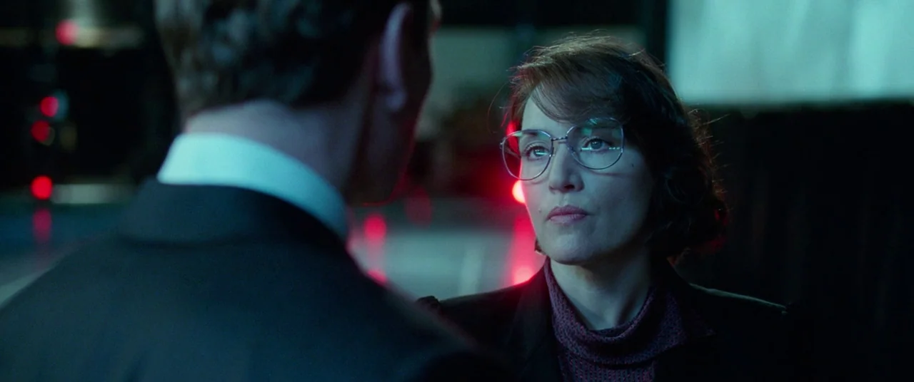 Steve Jobs (2015) movie still: medium — A dark, dimly lit indoor space featuring out-of-focus red glowing points in the background. A woman wearing…