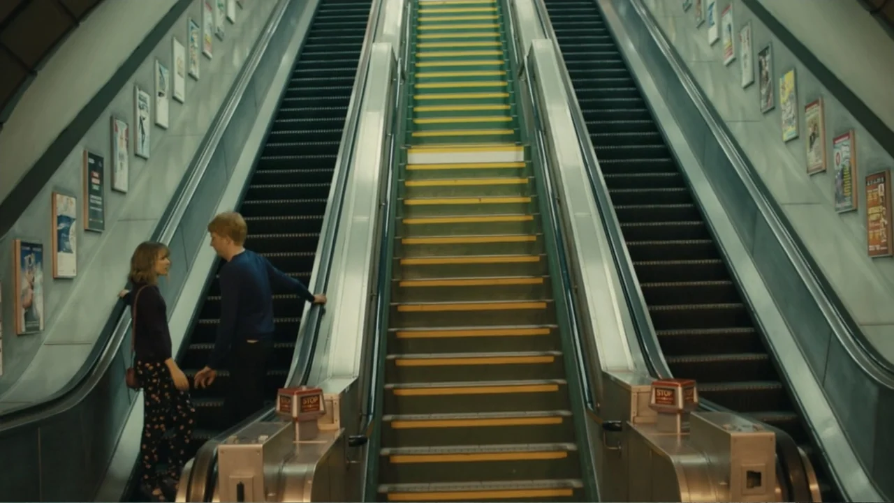 About Time (2013) movie still: wide — A long subway station escalator is flanked by walls covered in numerous rectangular advertisements. A young…