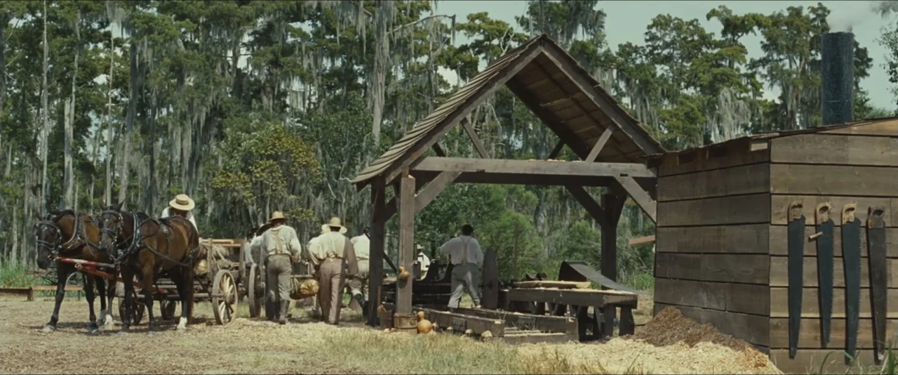 12 Years a Slave (2013) movie still: wide — An open-air wooden sawmill sits in a clearing surrounded by Spanish moss-draped trees. A group of men in…