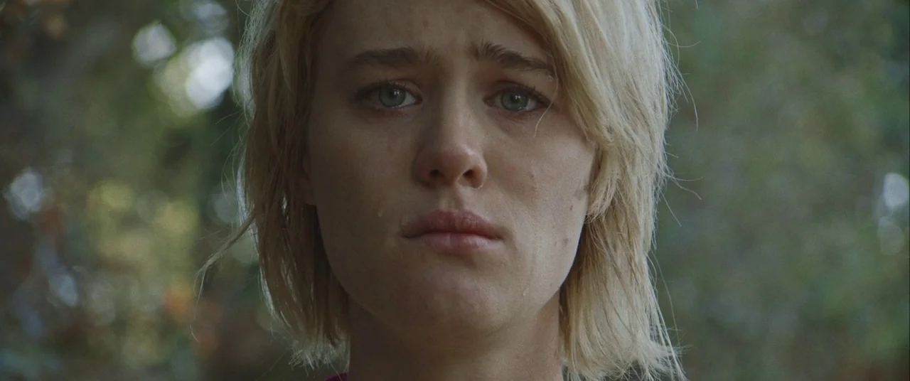 Always Shine (2016) movie still: closeup — A woman stands in a blurred outdoor environment with foliage in the background. She looks directly at the…