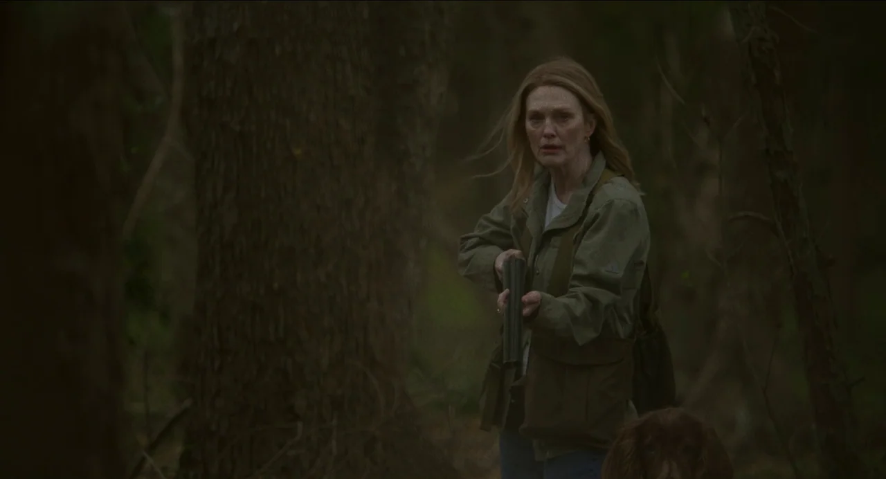 May December (2023) movie still: medium — A dense patch of dark, overgrown woodland with thick tree trunks and leaf litter. A woman in an olive…