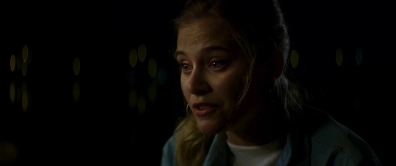 Summer of 84 (2018) movie still: closeup — A dark outdoor setting at night features soft bokeh lights in the distance. A young woman speaks while…