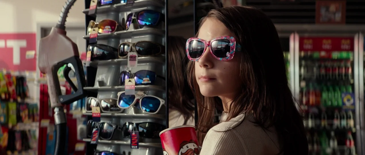 Logan (2017) movie still: medium — A young girl stands in a brightly lit retail shop aisle beside a rack. She holds a cylindrical snack container…