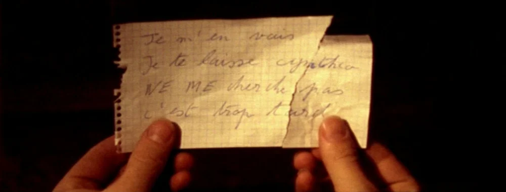 Carne (1991) movie still: closeup — Hands hold a torn piece of white grid-lined paper against a dark background. The paper features handwritten…