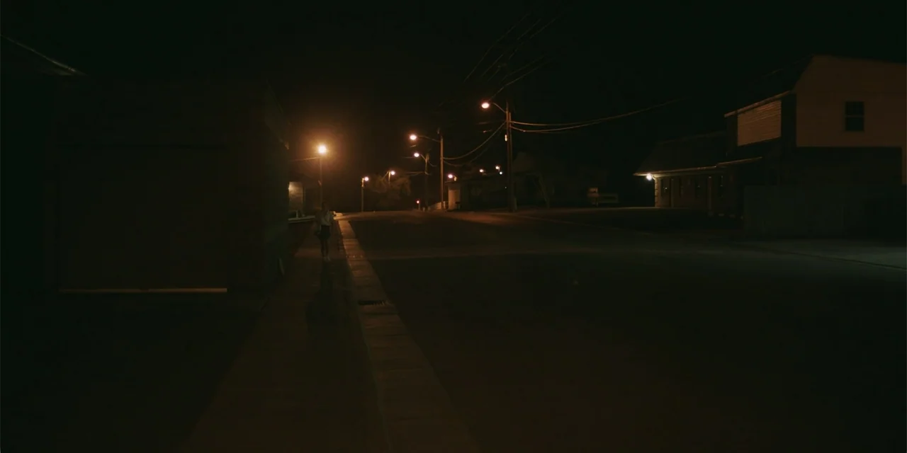 La coda del diavolo (2024) movie still: wide — A quiet suburban road at night is lit by spaced-out sodium-vapor street lamps. A lone individual…