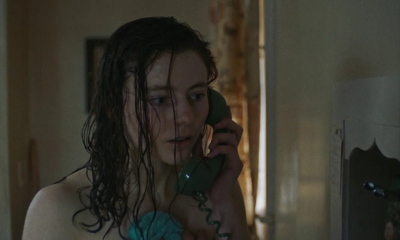 Eileen (2023) movie still: closeup — A dimly lit room with muted wall colors features a vintage wall-mounted telephone. A young woman with wet hair…