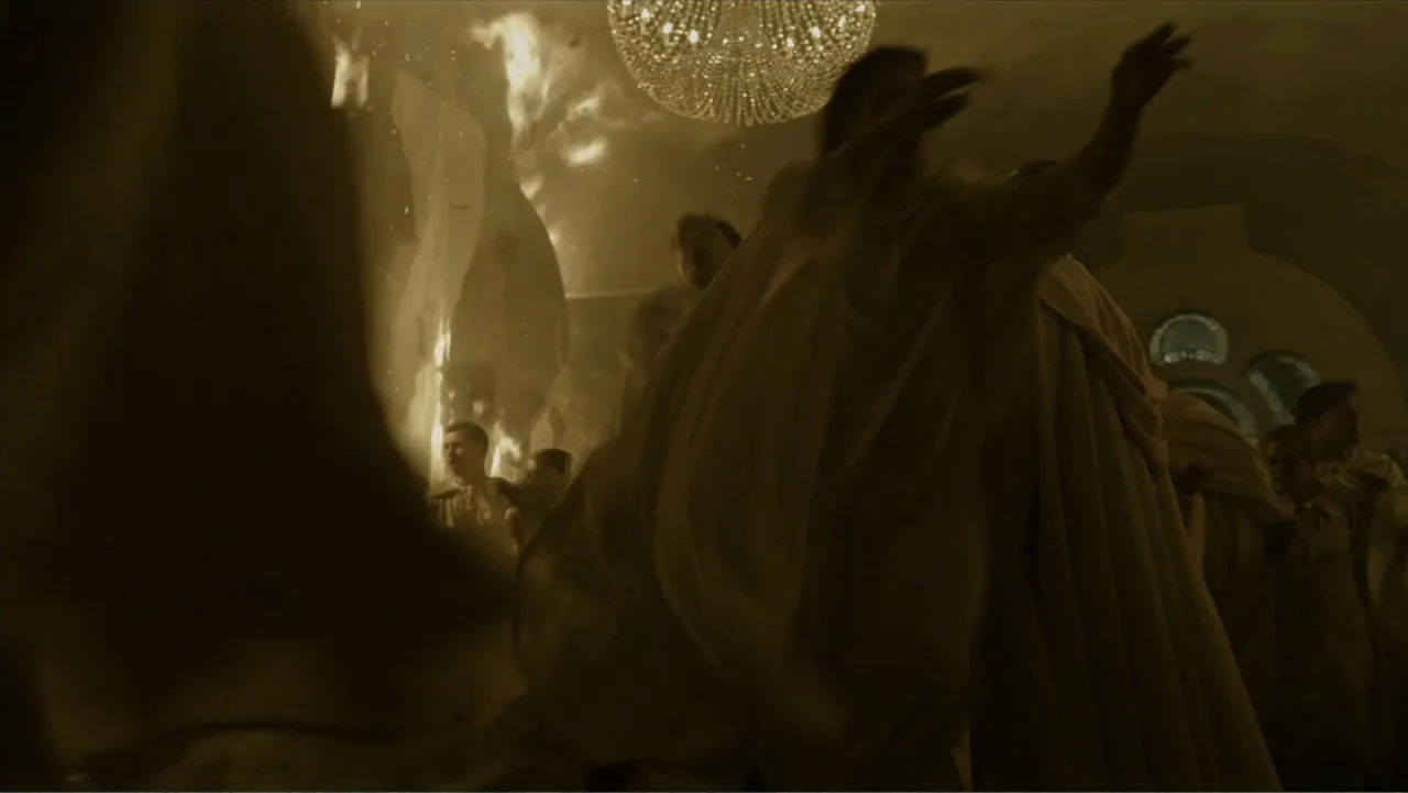 A Cure for Wellness (2017) movie still: medium — A stone interior space features a large chandelier and rising flames. People in draped fabric…