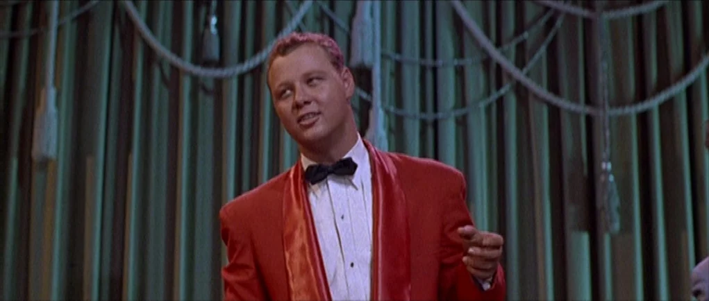 The Girl Can’t Help It (1956) movie still: medium — A man in a red formal jacket stands before a gathered teal curtain. He is speaking directly to…