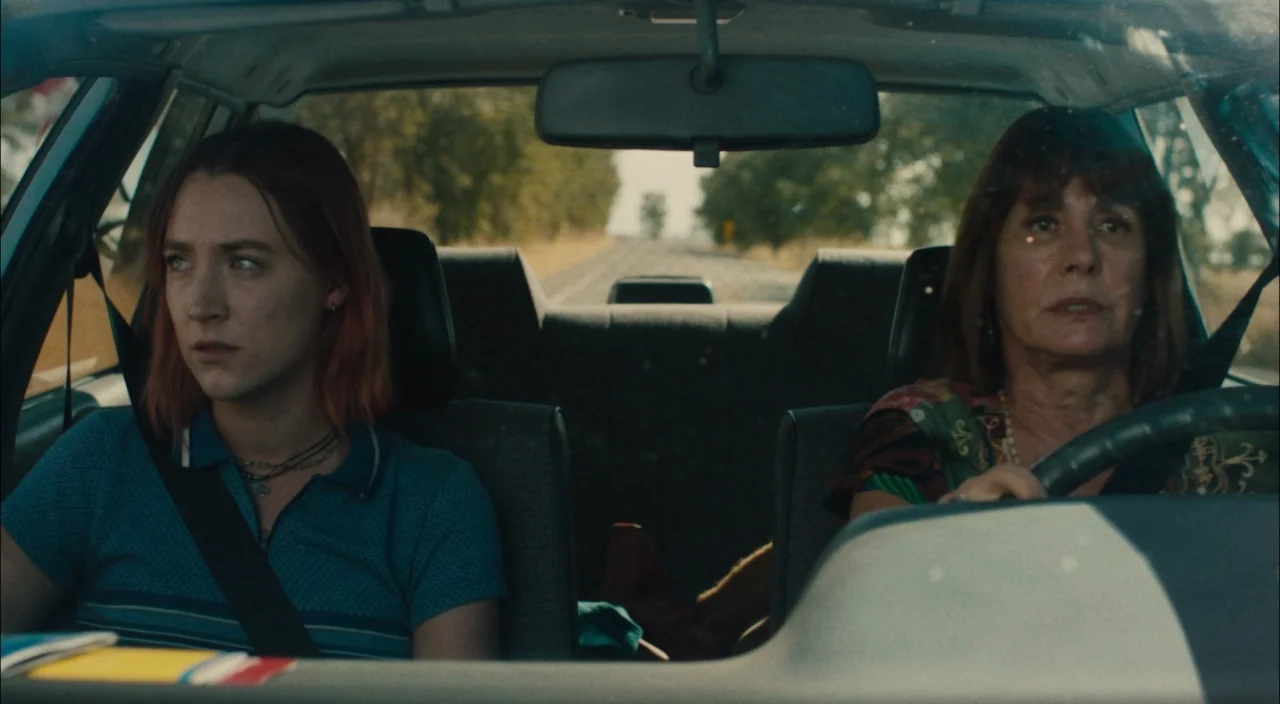 Lady Bird (2017) movie still: medium — A view from the backseat of a car onto the front seats and windshield. A young woman and an older woman are…