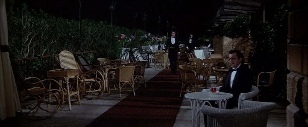 Death In Venice (1971) movie still: wide — An outdoor restaurant terrace at night features wicker furniture and a central red carpet. A man in a…