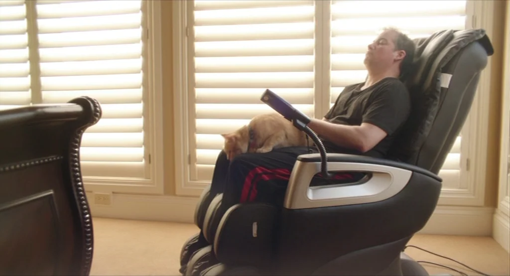 Results (2015) movie still: medium — A man sits in a black reclining chair with a orange cat on his lap. Bright light fills the room through the…