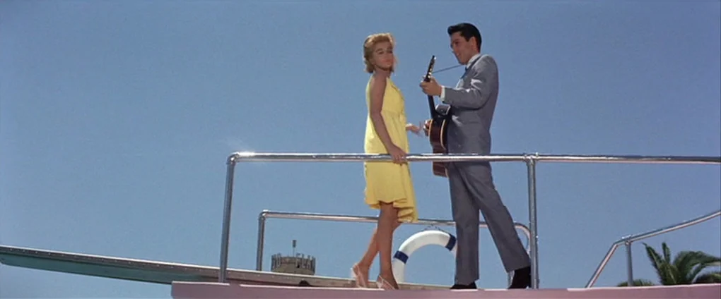 Viva Las Vegas (1964) movie still: fullbody — A sunny yacht deck against a clear blue sky. A man in a suit plays guitar for a woman in a yellow dress.