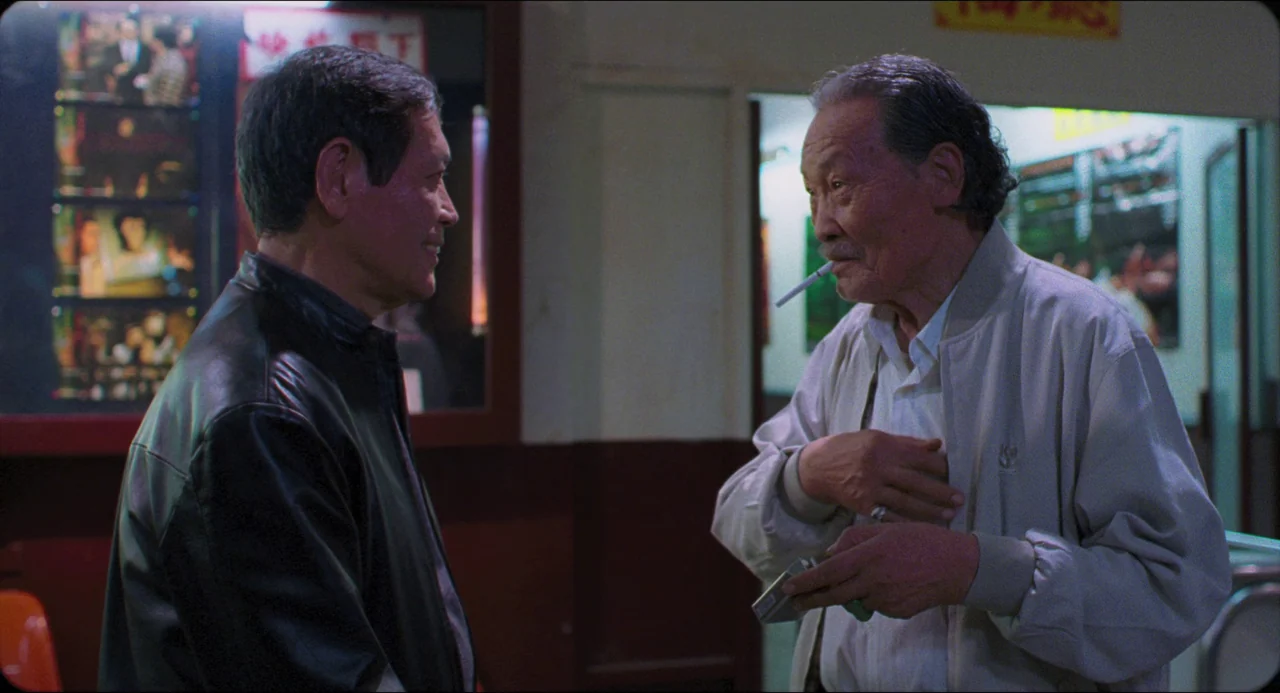 Goodbye, Dragon Inn (2003) movie still: medium — Two men converse in a dimly lit public area featuring a film display wall. The older man holds a…