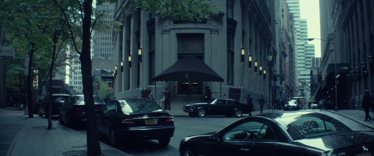 John Wick (2014) movie still: establishing — A stone building with a dark awning stands on a New York City corner. Several black cars are parked…