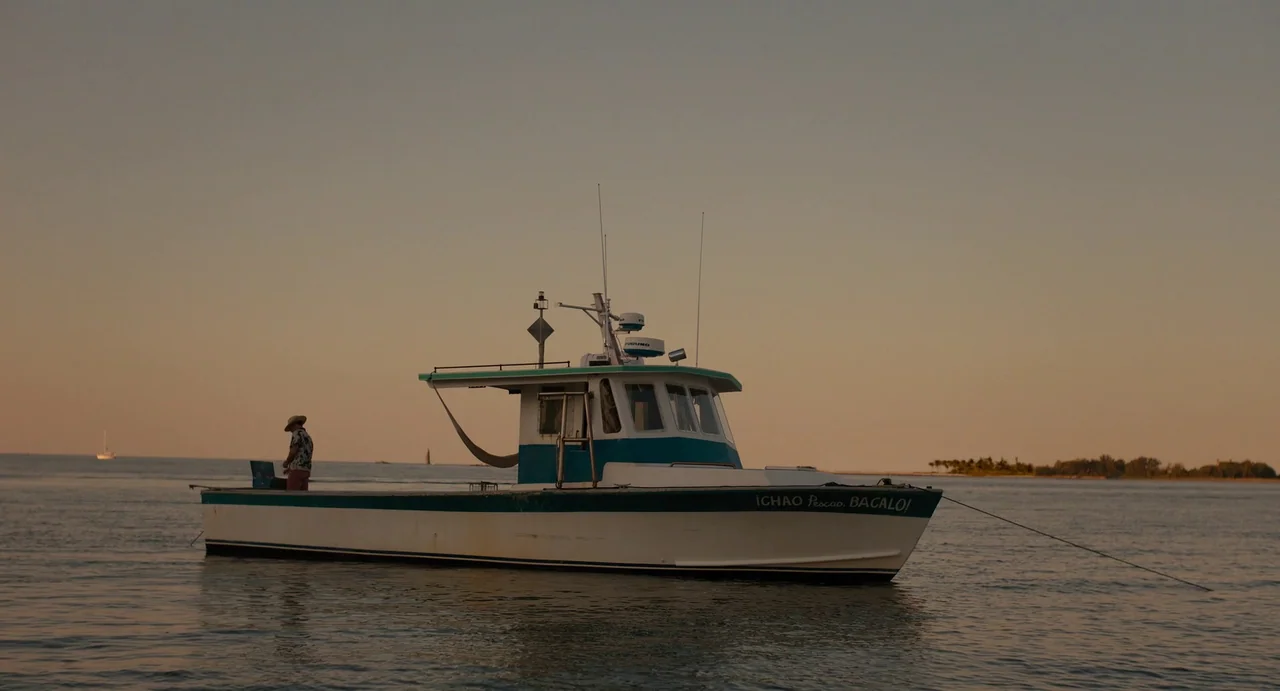 Confess, Fletch (2022) movie still: wide — A small white and teal motorboat floats on calm water during late afternoon. A single individual stands…