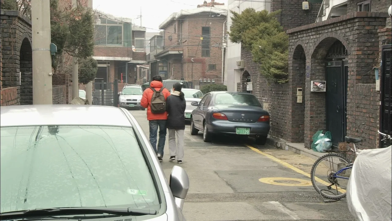 Oki’s Movie (2010) movie still: fullbody — A narrow residential street features brick buildings and parked cars under an overcast sky. Two…