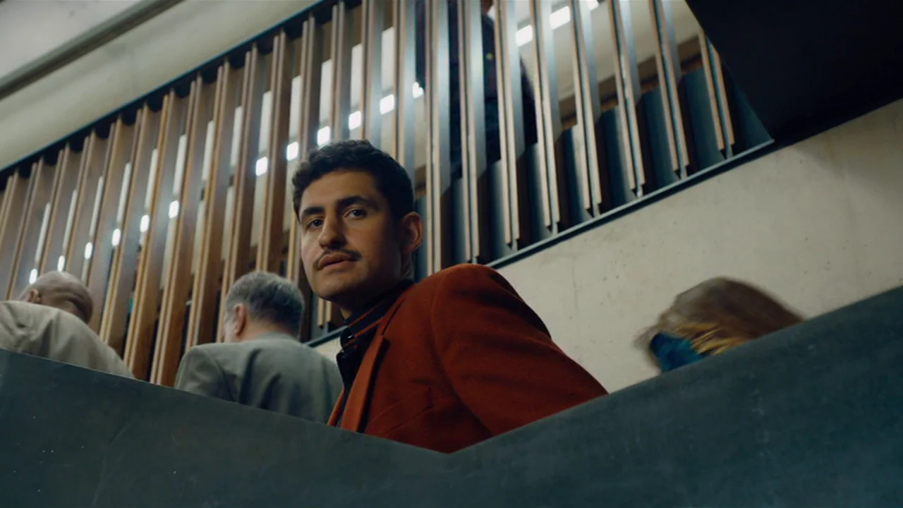 Club Zero (2023) movie still: medium — A man stands on a moving escalator inside a building with concrete walls. Behind him, wooden slats line the…