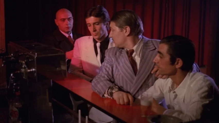 Tenderness of the Wolves (1973) movie still: medium — A dark, red-lit interior bar with men gathered along the counter surface. The men are sitting…